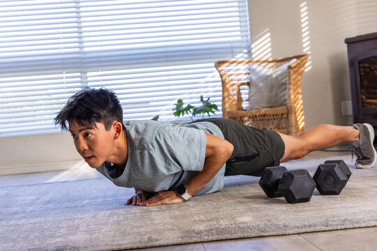 Asian man performing push-up on rug at home wearing gray sportswear with black hex dumbbells