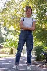Fototapeta premium Woman standing arms crossed, facing camera on park path in white tee, jeans, sunglasses