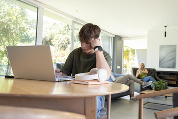 Obraz premium Family with teenage son at home sitting at table, using laptop and smartphone white headset nearby