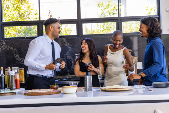 Four friends in business-casual attire gathering at kitchen island serving pizzas, holding drinks