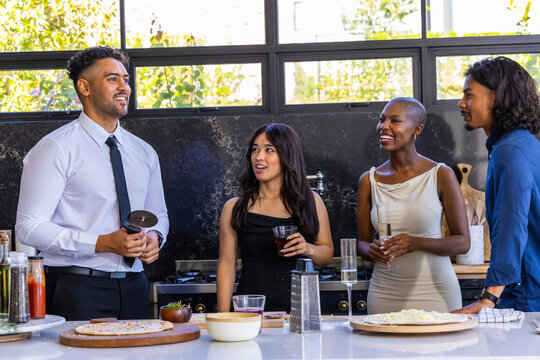 Diverse friends preparing pizzas on kitchen island, one man in shirt and tie using pizza cutter