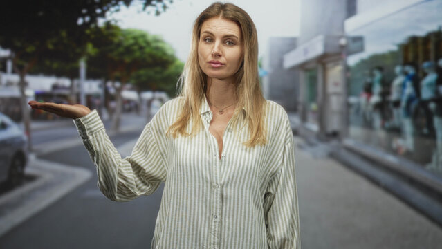 Woman with left palm up in a shrug gesture on a street storefront sidewalk, wearing a striped shirt and necklace, indifferent skeptical expression; skepticism.