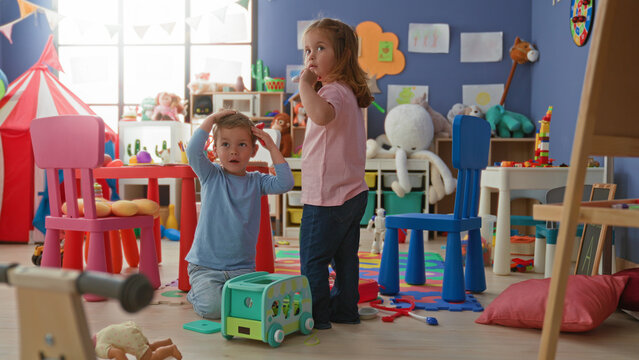 Girl and boy playing with a toy bus, boy seated touching his cheek while girl reaches for blocks amid colorful toys in building playroom; playful curiosity.