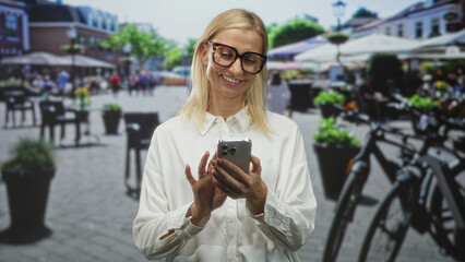 Fototapeta premium Woman holding smartphone, tapping screen while smiling in street cafe with bicycles and planters; happiness connection ease.