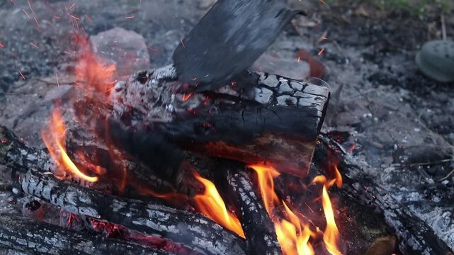The process of raking coals in a bonfire with a metal shovel. Close-up of burning firewood, ash and sparks. A metal shovel rakes up bright red hot coals in an evening bonfire. Bright flame