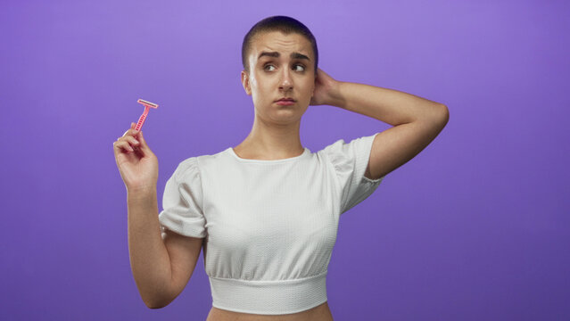 Woman with buzz cut holding pink disposable razor and hand on head in studio with purple backdrop wearing white crop top and puzzled expression; hesitation doubt.