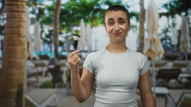 Woman with short hair holds makeup brush up near face with a half shrug expression in building; playful confusion.