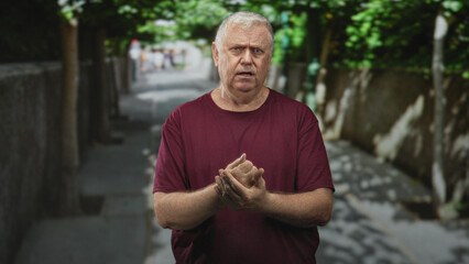 Fototapeta premium Man massaging and rubbing his hands on a shaded street path beside a stone wall while wearing a maroon tshirt; aging solitude discomfort.