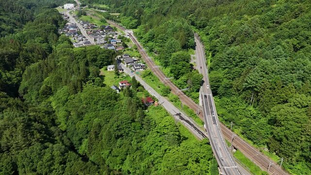 Aerial view above a winding road and railway track. Vibrant green forests surround the infrastructure. Small village lies nestled in the valley below. No people visible, so emotions not applicable