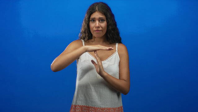 Hispanic woman in white tanktop making timeout sign with hands in studio with blue backdrop and soft lighting, embroidered hem visible; calm reflection.