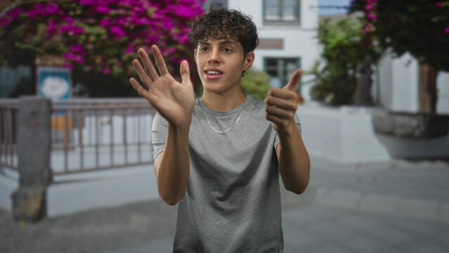 Teenage man lifts open hand to block his face while miming steering with the other hand in street near flowering bushes and railing; playful energy.