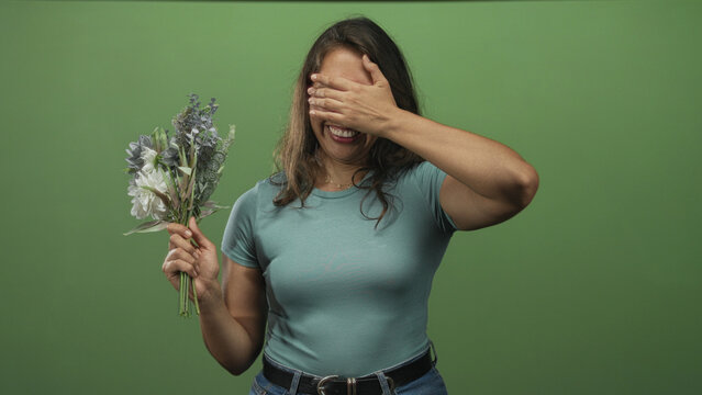 Woman holding bouquet of white and muted blooms, covering her eyes with one hand in green studio; shyness.