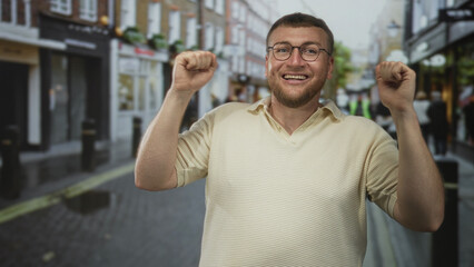 Naklejka premium Man smiling and fist pumping amid pedestrians on a busy urban street with shops and bollards; simple joy.