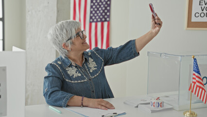 Woman takes selfie with smartphone and raised arm at polling building beside clear ballot box and...