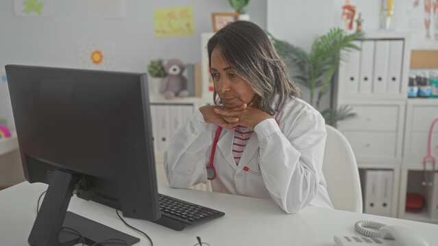 Hispanic woman doctor in white coat with stethoscope rests chin on folded hands at desk in building; concern.