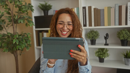 Woman with glasses holding tablet and smiling, showing hands and red nails in an office building with bookshelves and potted plants  remote work joy. © Krakenimages.com
