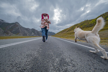 Low angle rear view of young woman with pink hair running with her white dog on empty asphalt road. Active female traveler with husky exploring mountain valley under cloudy sky. © ANDREY