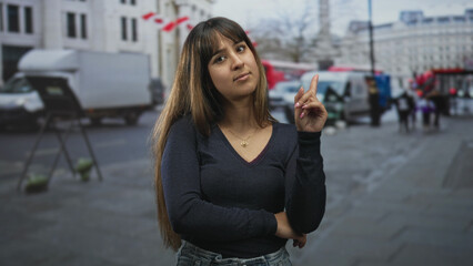 Fototapeta premium Young hispanic woman points index finger upward with folded arm and visible necklace on street near parked vans and pedestrians; quiet contemplation.