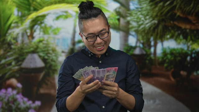Man counting indonesian rupiah banknotes with hands in forest setting, looking down; savings prosperity contentment.