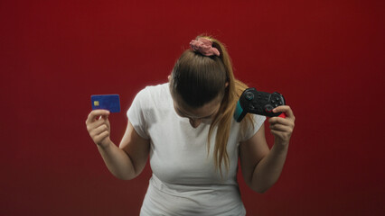 Woman holds a gamepad in her right hand and a credit card in her left while smiling and squinting, wearing a white t shirt in a red studio  excitement gaming. © Krakenimages.com