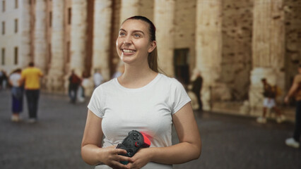 Woman holding a gamepad with both hands smiles and gazes upward amid a crowded plaza in front of a stone colonnaded building  playful joy. © Krakenimages.com