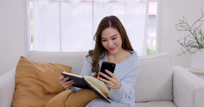 Young Asian woman relaxes on sofa, checks smartphone, writes activity note in planner, moves pen across page, enjoying productive home lifestyle and calm weekend organization in bright room corner
