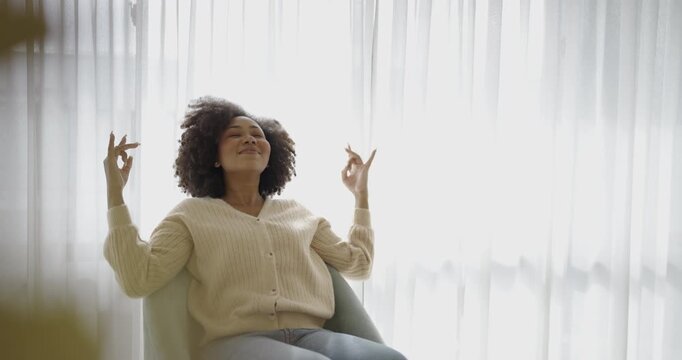 Smiling woman sitting indoors with hands in meditation gesture, expressing mindfulness, relaxation, mental wellness, and a calm lifestyle.