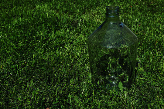 A large empty glass carboy or demijohn bottle standing outdoors on a green lawn in sunlight. The wide composition features an expansive grass texture on the left side, ideal for winemaking, home brewi