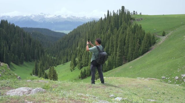 Travel mountain valley forest meadow hiker backpack smartphone snow pine man capturing landscape on alpine slope green hills conifer trees distant peaks summer adventure photography under cloudy sky