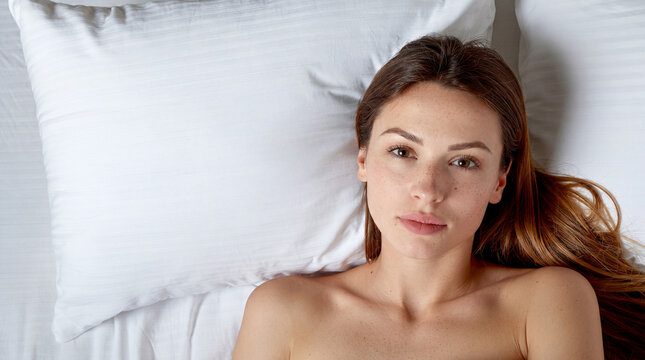 Beautiful young woman lying in bed on white pillows looking at camera