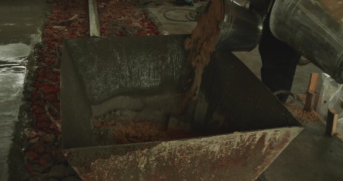 Unrecognizable male builder in workwear pouring sand from a bucket into a mixing container to prepare fresh concrete mortar for flooring at an indoor construction site