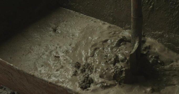 Professional construction worker using a shovel to manually mix wet cement mortar in a container, preparing a concrete mixture for floor screed or foundation work on a building site