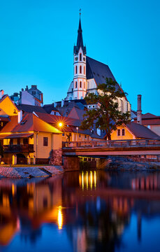 Czech Krumlov, Czech Republic. Evening view at bridge in old town with old houses and broach at tower of St. Vitus Church. Sunset blue hour with evening illumination.