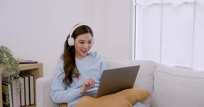 Young Asian woman wears headphone while working on laptop, listening music, tapping keyboard, holding cup, moving gently to rhythm for stress relief in cozy bright apartment home office routine
