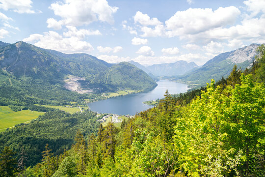 Panoramic view of Lake Grundlsee from the Lustiges Eck viewpoint in the Salzkammergut region of Styria, Austria. Wooden bench overlooking the alpine landscape with mountains, forest, and blue sky
