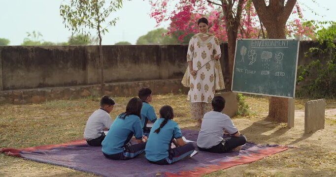 Indian young adult female tutor hold stick pointing words teach to girl boy rural area place. Village group sitting on mat look board talk speak read do study learn clap hands open ground class field