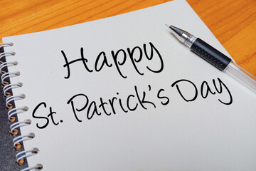 Obraz premium Top view of an open spiral notebook with Happy St. Patrick's Day handwritten text overlay next to a black gel pen on a wooden desk