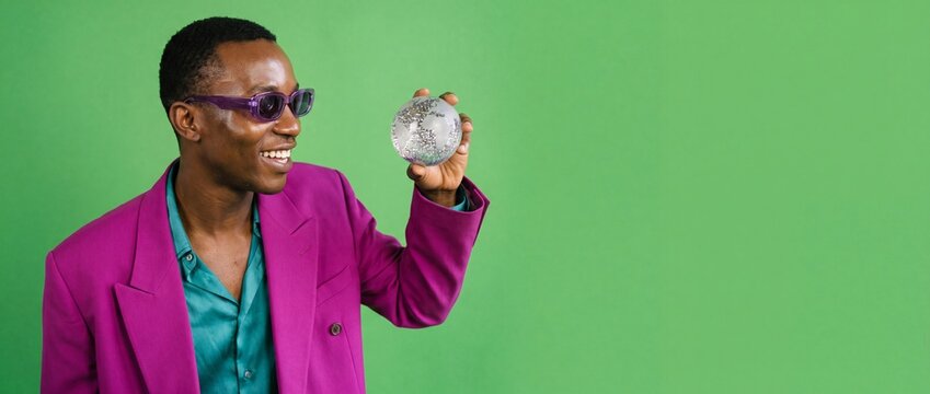 Black man holding silver globe in hand wearing magenta jacket and sunglasses isolated on green background, smiling with joy representing global business success and eco friendly vision banner concept.