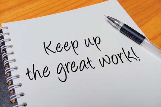 Top view of an open spiral notebook with Keep up the great work! handwritten text overlay next to a black gel pen on a wooden desk