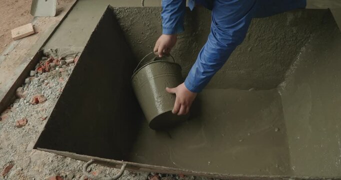Male builder scooping wet cement from a container with a bucket and pouring the mixture onto the floor during renovation work, creating a smooth and level screed for a new surface