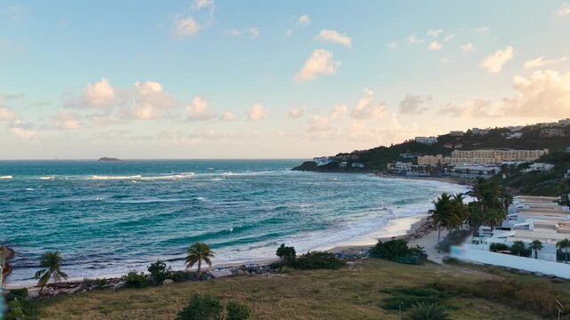 Aerial view of Dawn Beach on the Dutch side of Saint Martin island in the Caribbean. Turquoise ocean waves roll onto the white sand coastline. Low-rise tropical villas and green hills line.