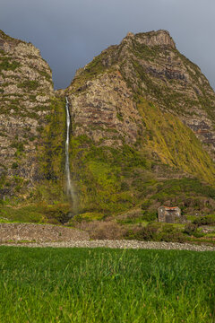 Impresionante paisaje de una alta cascada que fluye desde un acantilado rocoso y verde, con un prado en primer plano y una casa r&uacute;stica de piedra en la pintoresca isla de Flores, Portugal.