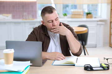 Naklejka premium Workaholic. Overwhelmed man yawning at wooden desk with laptop, takeaway cup of coffee and notebook in kitchen