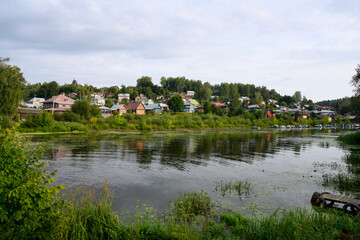 Fototapeta premium A river backwater covered with reeds and water lilies, with wooden houses, boats and a church in the town of Ples, Russia