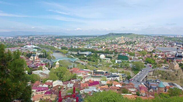 Panoramic view of Tbilisi, Georgia, captured from an elevated viewing platform. The footage shows the cable car gliding above the Mtkvari River canyon, Metekhi Church on the cliff edge, and Rike Park.