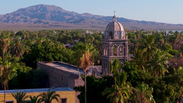 Watch sunrise aerial views of Loreto Mission church in Baja California Sur, Mexico as it shines over the landscape and palm trees.