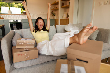 Plakat Excited woman sitting on sofa surrounded by many delivery parcels and raising hands in winner gesture, celebrating arrival of online shopping orders