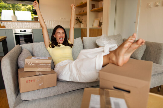 Excited woman sitting on sofa surrounded by many delivery parcels and raising hands in winner gesture, celebrating arrival of online shopping orders