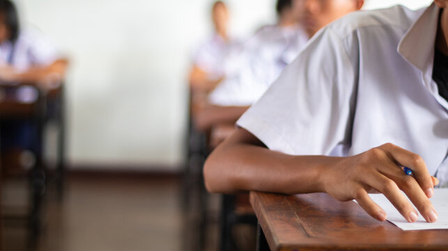 Close-up of a student's hand diligently writing on an exam paper during a school test. 
The student is using a professional pen , sitting at a wooden desk in a brightly lit classroom.