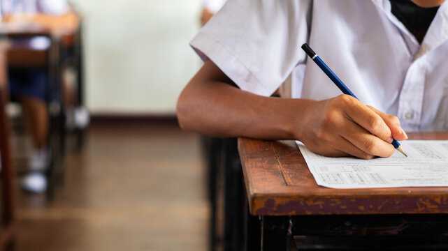 Close-up of a student's hand diligently writing on an exam paper during a school test. 
The student is using a professional pen , sitting at a wooden desk in a brightly lit classroom.